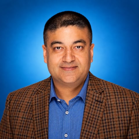 Professional headshot of a man wearing a brown plaid blazer over a blue checkered shirt against a blue background